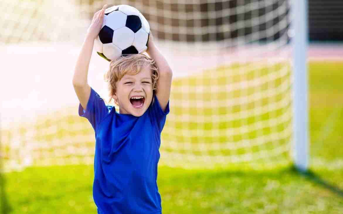 Benefícios do futebol infantil. Menino de blusa azul segurando bola em cima da cabeça e perto de um gol de futebol