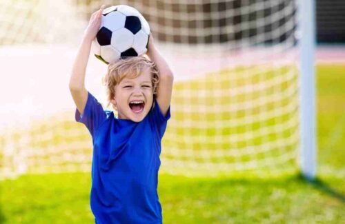 Benefícios do futebol infantil. Menino de blusa azul segurando bola em cima da cabeça e perto de um gol de futebol