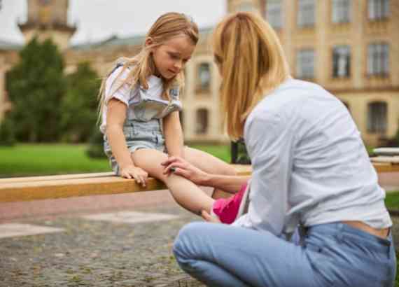 Como aliviar as dores de crescimento em crianças e adolescentes. Criança sentada em um banco da praça com uma mulher examinando sua perna que doí