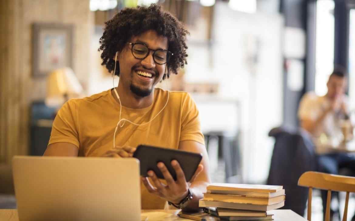 Homem sorrindo, de fone de ouvido, segurando um celular na mão e de frente a um notebook e uma pequena pilha de livros ao seu lado. O lado bom da internet.