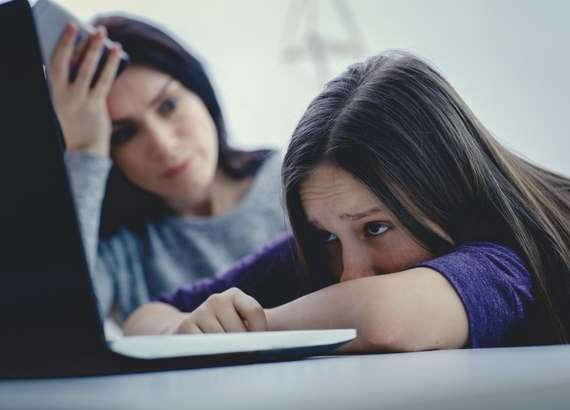 Os efeitos do TikTok em crianças e adolescentes - menina pré-adolescente olhando para a tela do notebook com feição muito triste e mãe, ao fundo, observa-a preocupada. 