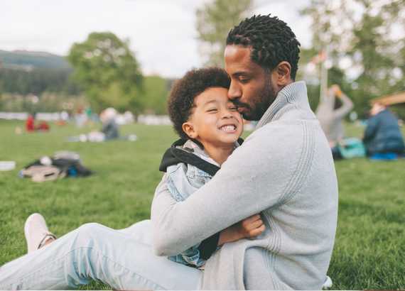 Homem sentado no gramado de um parque com seu filho pequeno no colo. A importância da licença paternidade para a igualdade de gênero 
