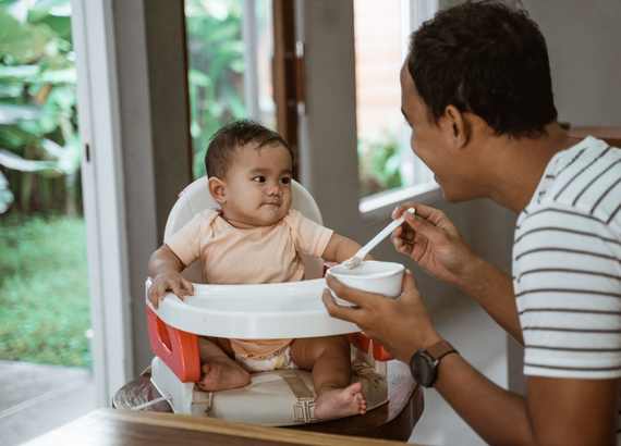 Criança em uma cadeirinha de alimentação e o pai lhe dando comida. A importância da licença paternidade para a igualdade de gênero 