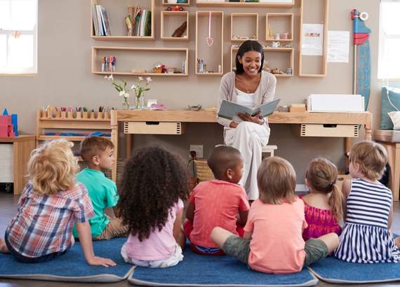 Professora lendo para a classe de pequenas crianças sentadas no chão sobre almofadas. - Como ensinar a ler 8 dicas fáceis e práticas 