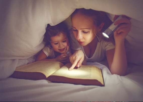 Mãe e criança pequena sob uma cabana feita de cobertores sobre a cama. Mãe segura uma lanterna. - Como ensinar a ler 8 dicas fáceis e práticas 
