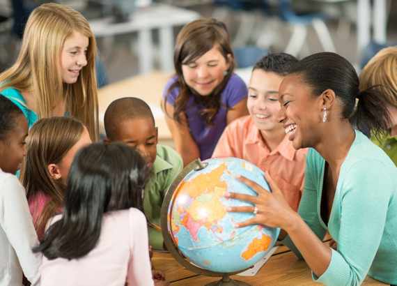 Como escolher um método de ensino para o meu filho? Professora segurando um globo terrestres cercada por seus alunos.