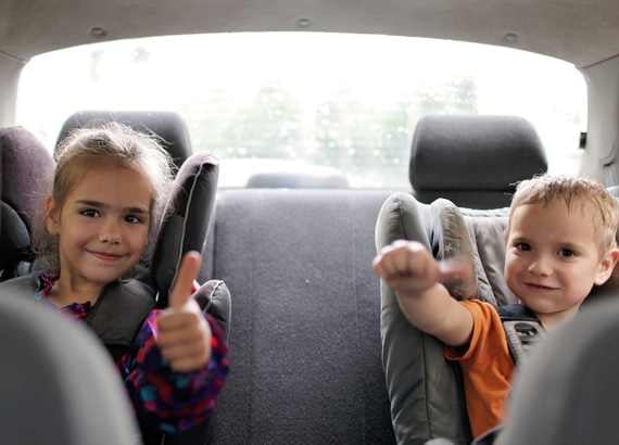 Duas crianças no banco de trás do carro, em cadeiras de segurança, fazendo sinal de positivo e sorrindo.