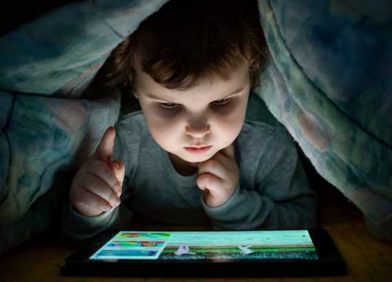 Criança debaixo das cobertas mexendo em tablet.