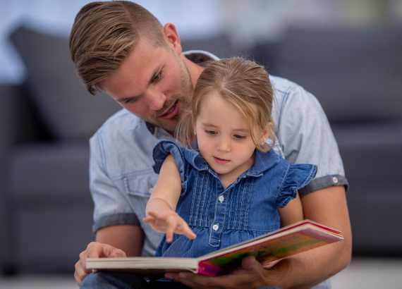 Pai com a filha no colo lendo um livro infantil juntos.