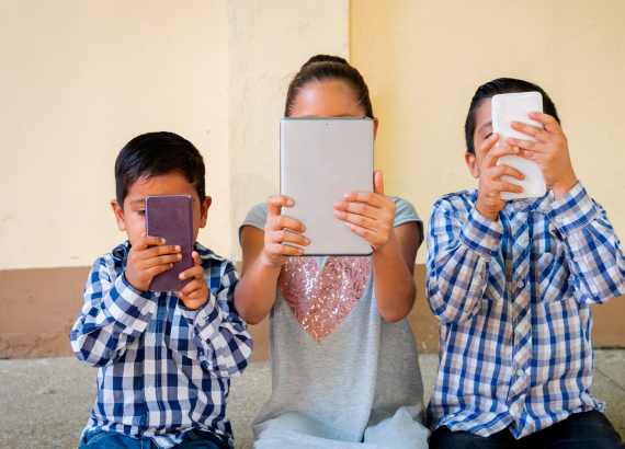 Três crianças com smartphones e tablets logo à frente de seus rostos.