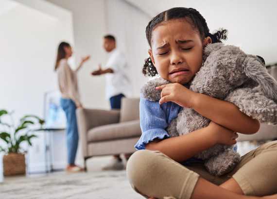 O que é a depressão infantil?
Menina com cara de choro, tristeza extrema, abraçada com um bicho de pelúcia enquanto ao fundo há uma discussão de casal.