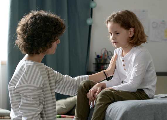 Por que é importante a criança aprender a lidar com a frustração ainda na infância. Mãe conversando com a filha que está sentada em um sofá. A mãe está ajoelhada em sua frente.