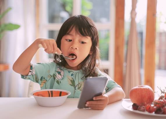 Excesso de telas: como a mente infantil reage a tantos estímulos? 3 Telas em excesso como a mente infantil reage a tantos estímulos_meio. Menina comendo com celular na mão