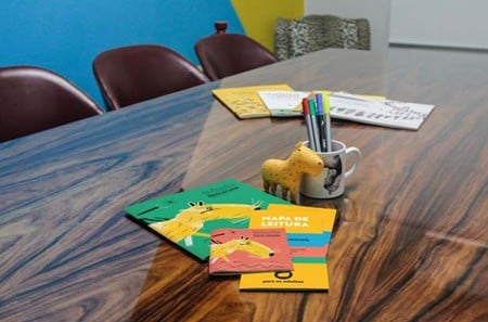 Foto de uma mesa de reuniões de madeira com mapas de leitura do Clube Quindim, uma caneca com canetas e um mascote de pelúcia da Capivara Quindim